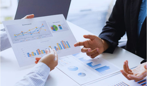 Small business growth consultant meeting with a client at a desk, reviewing printed business charts and performance data to discuss strategies for company growth.